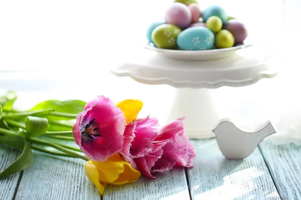 Ovos de Páscoa em vaso e tulipas na mesa em fundo brilhante — Fotografia de Stock