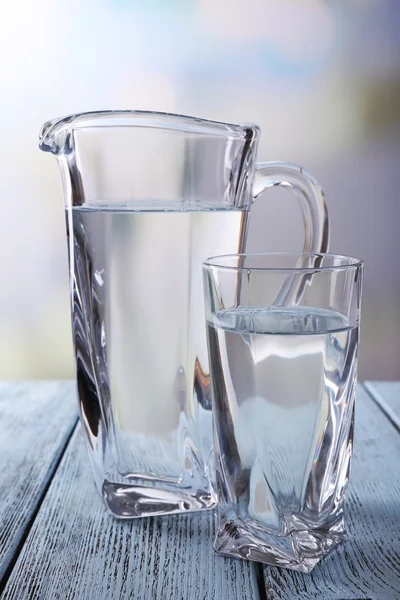 Brocca di vetro e bicchiere d'acqua su tavolo di legno su sfondo luminoso — Foto Stock