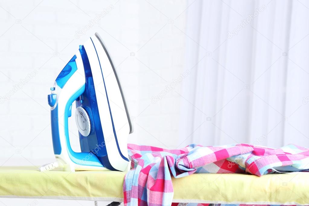 Iron and clothes on ironing board on interior background