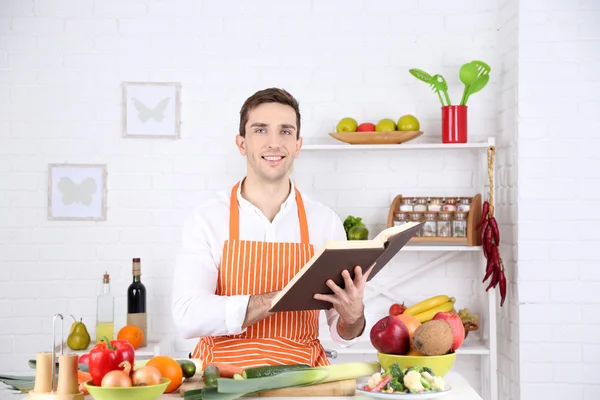 Человек с книгой рецептов в руках за столом с различными продуктами и посудой на кухне на белом фоне стены — стоковое фото