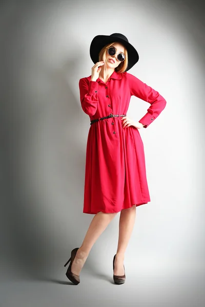 Expressive young model in red dress, black hat and sunglasses on gray background — Stock Photo, Image