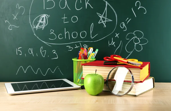 Digital tablet, books, colorful pens and apple on desk in front of blackboard — Stock Photo, Image