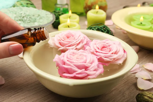 Vrouwelijke hand met fles essentie en kom met spa water met bloemen op houten tafel, close-up — Stockfoto