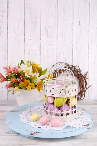 Easter composition with Easter eggs in decorative cage and flowers, on color wooden background — Stock Photo, Image