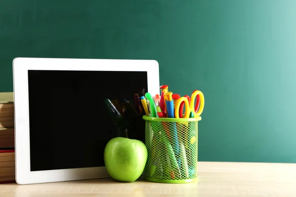 Digital tablet, books, colorful pens and apple on desk in front of blackboard — Stock Photo, Image