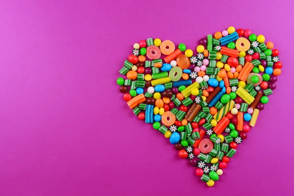 Colorful candies in heart shape on purple background — Stock Photo, Image