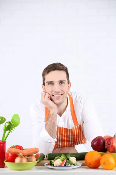 Mann am Tisch mit verschiedenen Produkten und Utensilien in der Küche auf weißem Wandhintergrund — Stockfoto