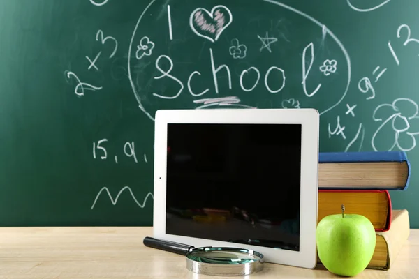 Digital tablet, books, colorful pens and apple on desk in front of blackboard