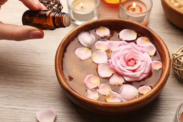 Female hand with bottle of essence and bowl of spa water with flowers on wooden table, closeup — Stock Photo, Image