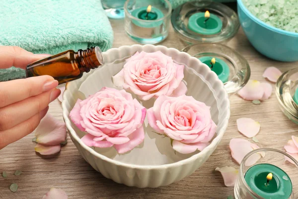 Female hand with bottle of essence and bowl of spa water with flowers on wooden table, closeup — Stock Photo, Image