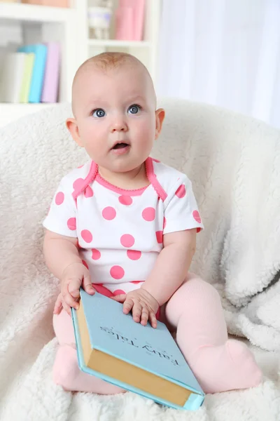 Cute baby girl sitting in arm-chair with book, on home interior background — Stock Photo, Image
