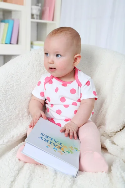 Söt baby flicka sitter i fåtölj med bok, på hem inredning bakgrund — Stockfoto