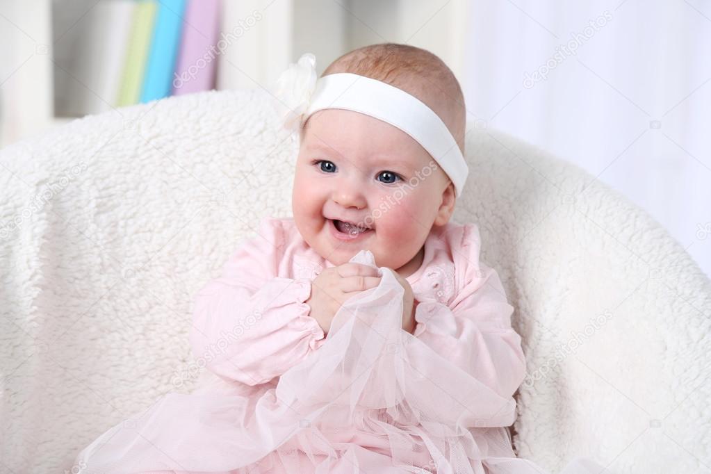 Cute baby girl in pink dress sitting in arm-chair, on home interior background