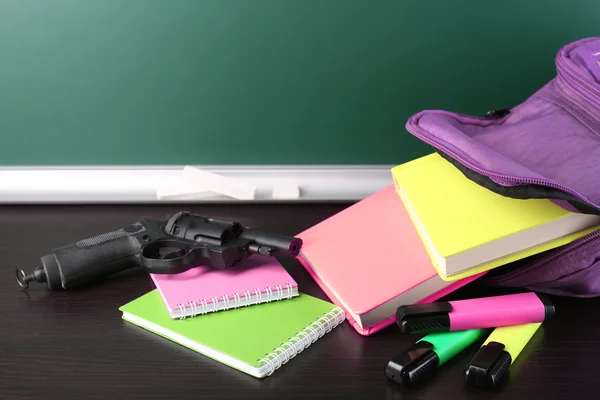 Pistola en la mochila de la escuela en escritorio de madera, sobre fondo de pizarra — Foto de Stock