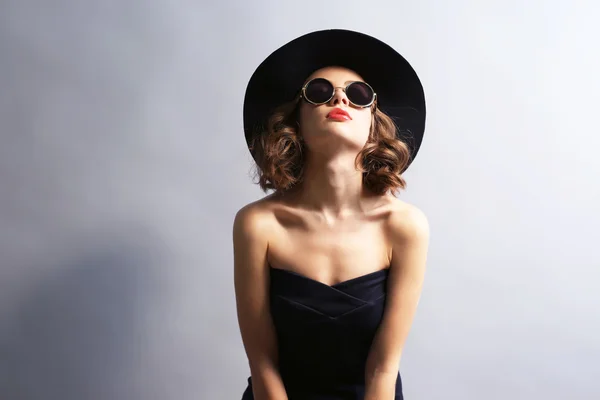 Retrato de modelo hermoso en vestido negro, sombrero y gafas de sol sobre fondo gris — Foto de Stock