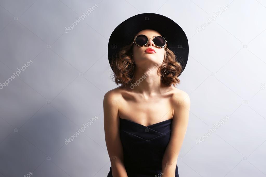 Portrait of beautiful model in black dress, hat and sunglasses on gray background