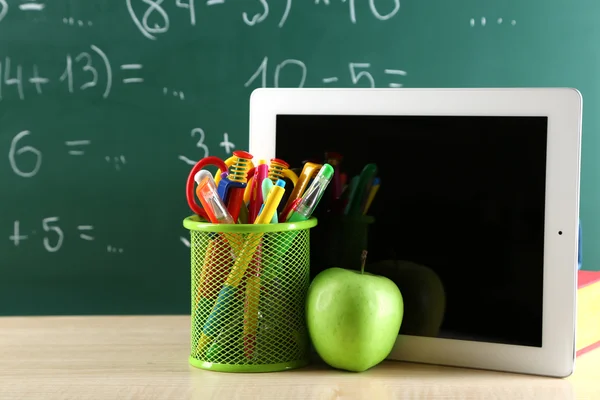 Tableta digital, libros, bolígrafos de colores y manzana en el escritorio frente a la pizarra —  Fotos de Stock