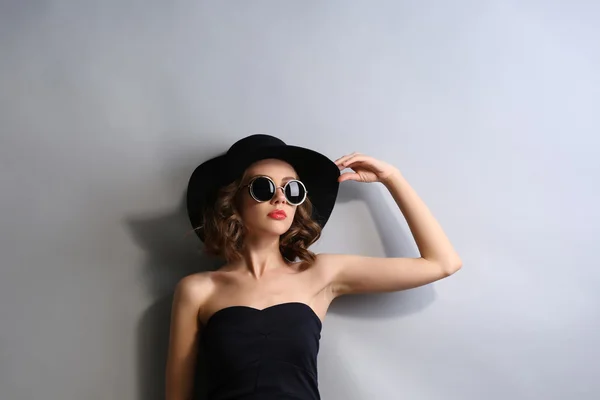 Retrato de modelo hermoso en vestido negro, sombrero y gafas de sol sobre fondo gris — Foto de Stock