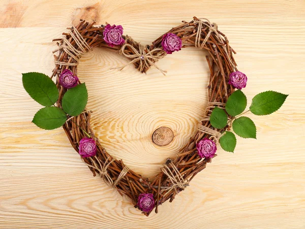 Wicker heart with dried roses and green leaves on wooden background — Stock Photo, Image