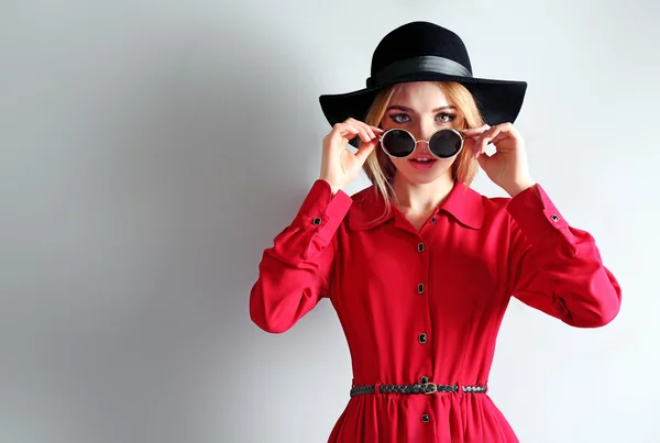 Экспрессивная молодая модель в красном платье, черной шляпе и солнечных очках на сером фоне — стоковое фото