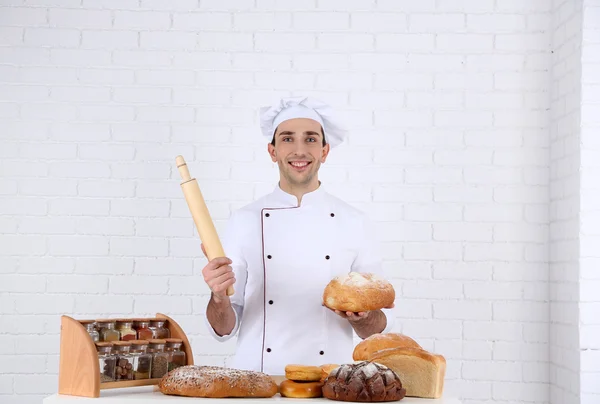 Piekarz w kuchni przy stole z świeżo bochenków chleba na biały cegła na ścianie tło — Zdjęcie stockowe