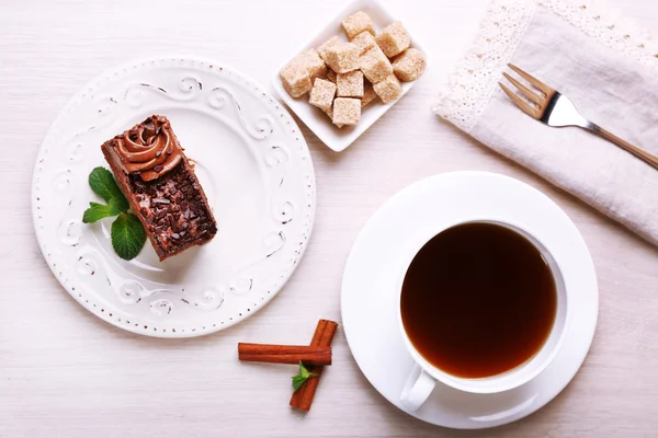 Sabroso pedazo de pastel de chocolate con menta y canela cerca de la taza de té y terrón de azúcar sobre fondo de mesa de madera — Foto de Stock