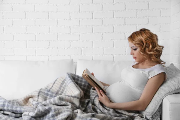 Junge schwangere Frau liegt auf Sofa und liest Buch auf hellem Hintergrund — Stockfoto