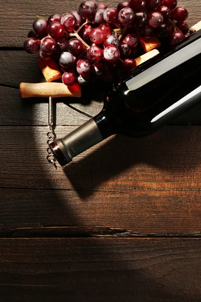 Glass bottle of wine with corkscrew and grapes in dark with light on wooden table background — Stock Photo, Image