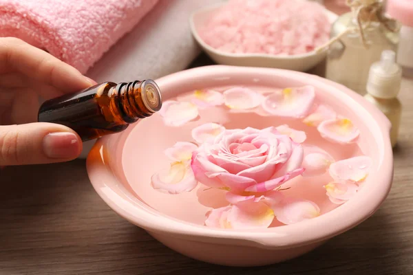 Tangan wanita dengan botol esensi dan semangkuk air spa dengan bunga-bunga di atas meja kayu, closeup — Stok Foto