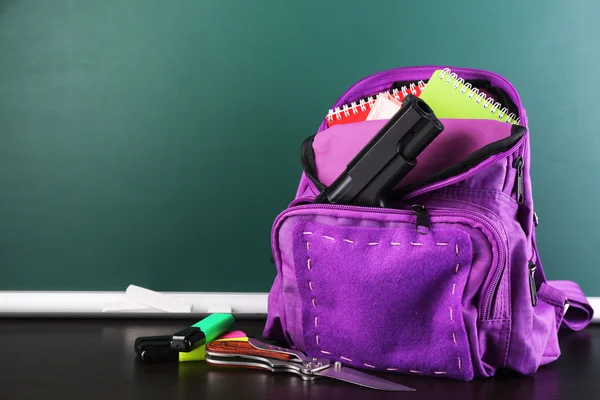 Pistola en la mochila de la escuela en escritorio de madera, sobre fondo de pizarra —  Fotos de Stock