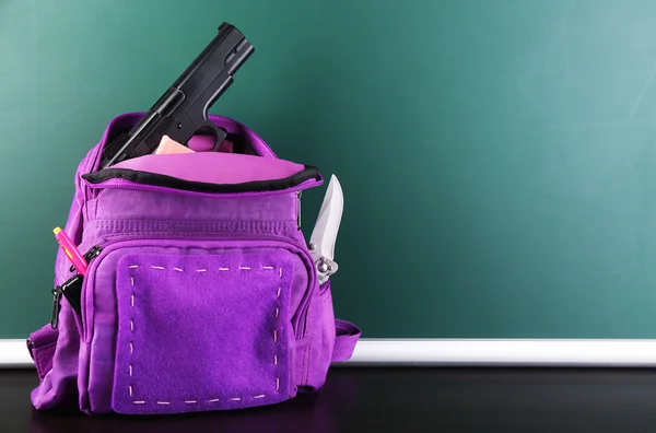 Pistola en la mochila de la escuela en escritorio de madera, sobre fondo de pizarra —  Fotos de Stock