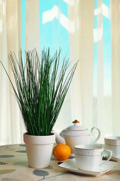 Green plant with teapot, cups and lemons on table on curtains background