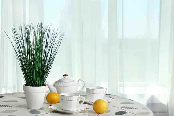 Planta verde con tetera, tazas y limones sobre mesa sobre fondo de cortinas — Foto de Stock