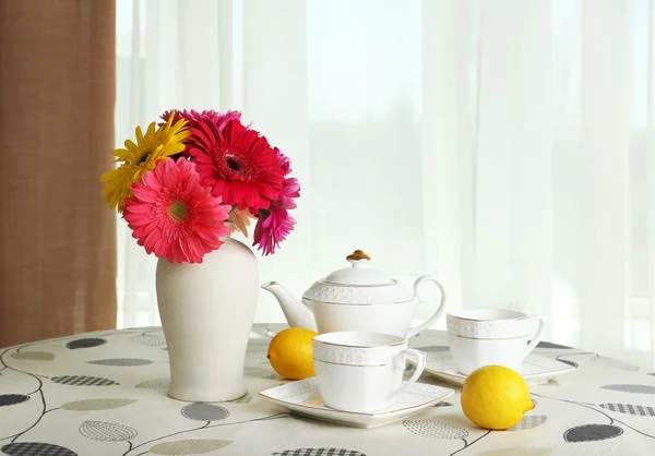 Colorful gerbera in vase with teapot, cups and lemons on table on curtains background — Stock Photo, Image