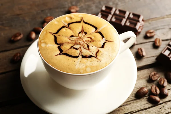 Cup of coffee latte art with grains and chocolate on wooden table, closeup — Stock Photo, Image