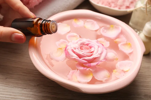 Mão feminina com garrafa de essência e tigela de água termal com flores na mesa de madeira, close-up — Fotografia de Stock