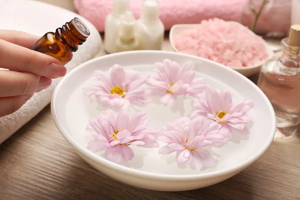 Mão feminina com garrafa de essência e tigela de água termal com flores na mesa de madeira, close-up — Fotografia de Stock