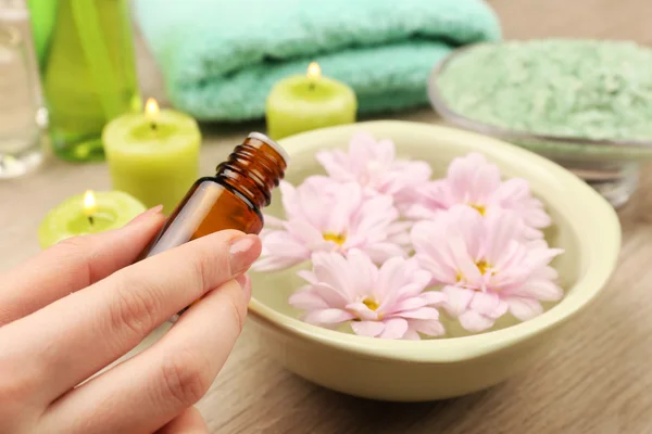 Female hand with bottle of essence and bowl of spa water with flowers on wooden table, closeup — Stock Photo, Image