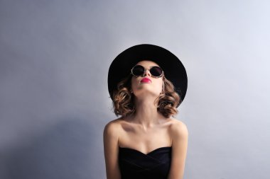Gri arka planda siyah elbise, şapka ve güneş gözlüğü takan güzel bir mankenin portresi.