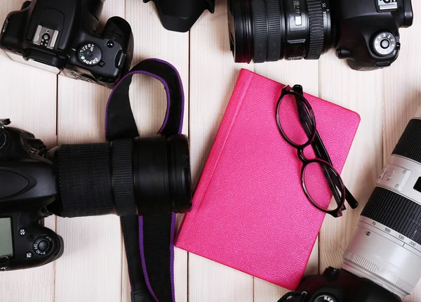 Modern cameras with glasses and pink diary on wooden table, top view — Stock Photo, Image