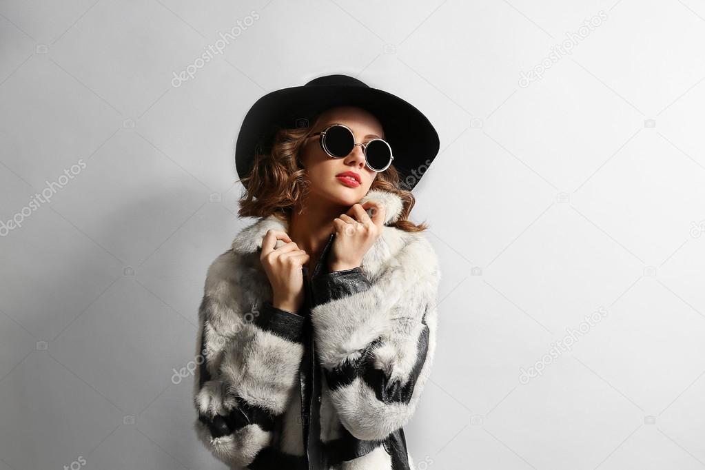Portrait of beautiful model in fur coat, hat and sunglasses on gray background