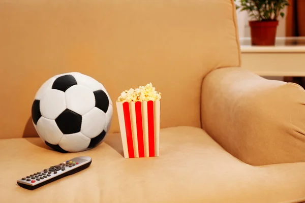 Soccer ball, remote control and box of popcorn on comfortable sofa, indoors — Stock Photo, Image