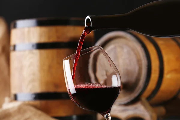 Pouring red wine from bottle into glass with wooden wine casks on background — Stock Photo, Image