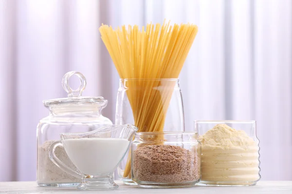 Pasta in glass bottle with cream and different types of flour on wooden table on curtain background