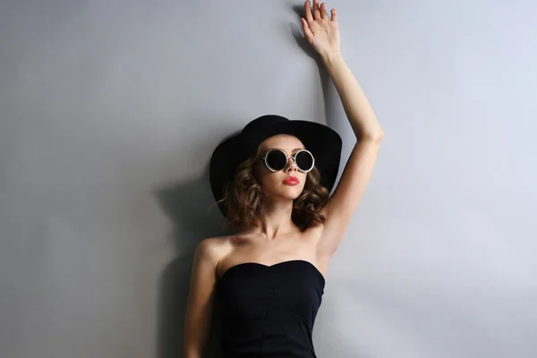 Retrato de modelo hermoso en vestido negro, sombrero y gafas de sol sobre fondo gris —  Fotos de Stock