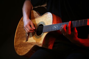 Genç adam akustik gitarla karanlık arka planda çalıyor.