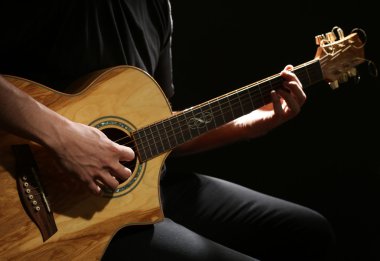 Genç adam akustik gitarla karanlık arka planda çalıyor.