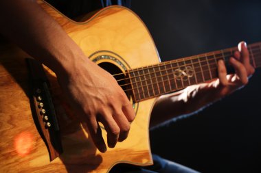Genç adam akustik gitarla karanlık arka planda çalıyor.
