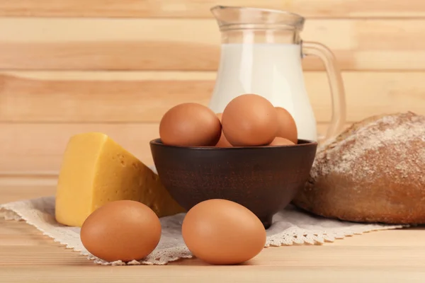 Huevos en tazón de barro, leche en jarra de vidrio, queso y pan sobre tabla de madera — Foto de Stock