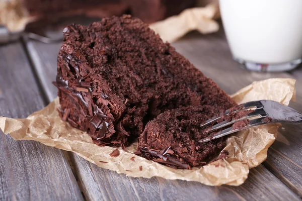 Pastel de chocolate sabroso en rodajas en hoja de pergamino y vaso de leche sobre fondo de mesa de madera —  Fotos de Stock